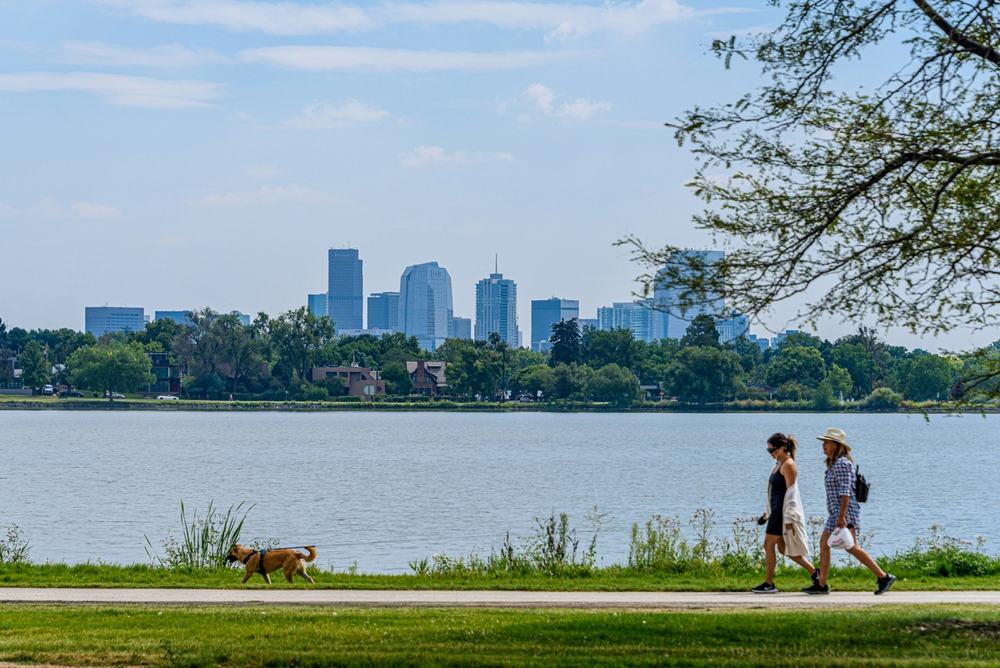 Two people walking along a lake with the Denver skyline behind